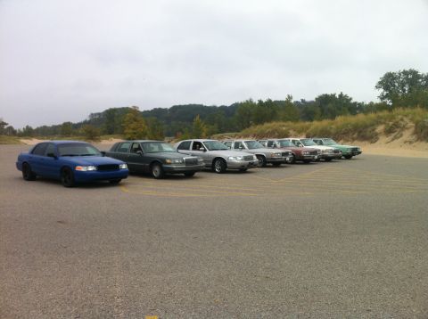 2003 Ford Crown Victoria - A line of sedans is parked in a lot with a sandy embankment and trees in the background. The closest vehicle is a blue sedan with black rim…