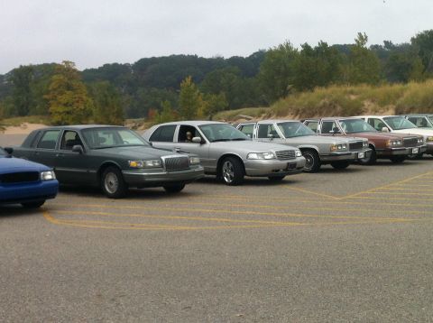 2003 Ford Crown Victoria - A line of sedans, including a dark green Lincoln Town Car and a silver Ford Crown Victoria, are parked in a lot with a wooded area and sand…
