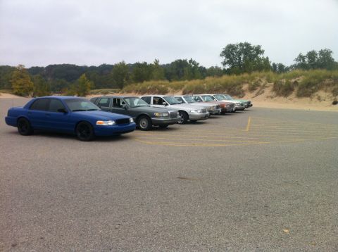 2003 Ford Crown Victoria - A line of sedans is parked on asphalt with yellow parking lines. The closest vehicle is a matte blue sedan with black wheels.
