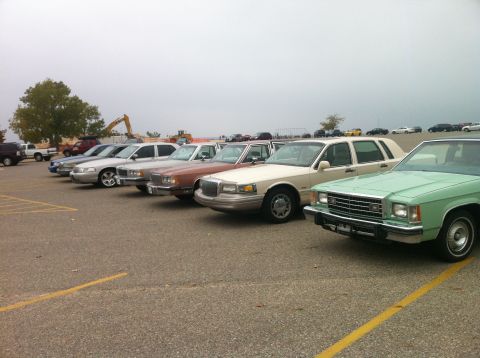 2003 Ford Crown Victoria - A line of sedans are parked in a lot under an overcast sky. The closest vehicle is a light green sedan with a chrome grille and bumper.