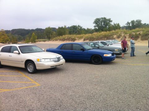 2003 Ford Crown Victoria - A line of sedans is parked in a lot with sand dunes and trees in the background. The closest vehicle is a cream-colored sedan, followed by…