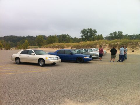 2003 Ford Crown Victoria - A line of sedans, including a light-colored Lincoln Town Car and a blue Ford Crown Victoria, are parked in a lot with a sandy dune and tree…