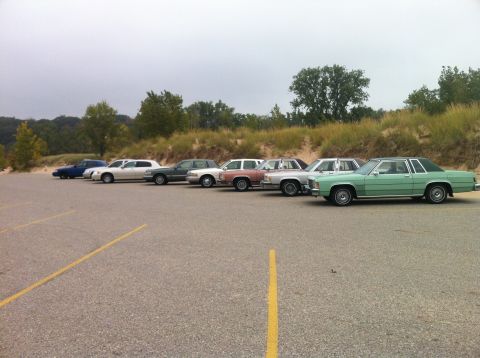 2003 Ford Crown Victoria - A line of vintage sedans are parked in a gravel lot with a grassy dune and trees in the background under an overcast sky. The vehicles are…