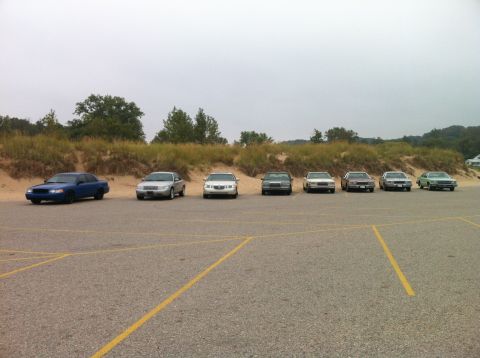 2003 Ford Crown Victoria - A line of sedans are parked in a lot with sand dunes and trees in the background. The closest vehicle is a blue sedan.
