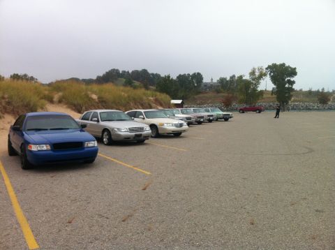 2003 Ford Crown Victoria - A line of sedans are parked in a lot with sand dunes and trees in the background. The closest vehicle is a blue Ford Crown Victoria, follow…