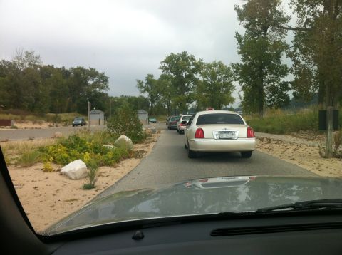 2003 Ford Crown Victoria - A line of cars, including a white Lincoln Town Car, are stopped on a paved road next to a sandy area with vegetation and trees under a clou…