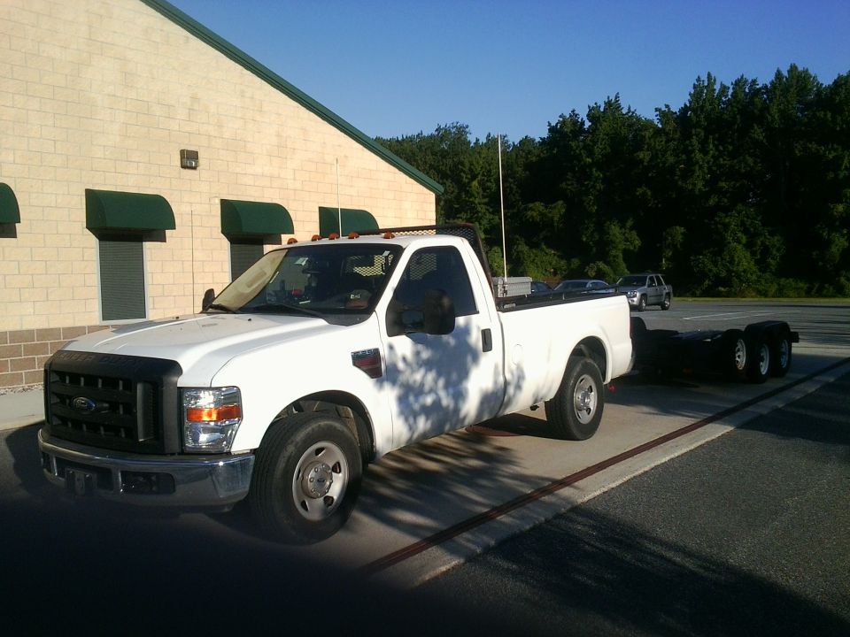 White Ford F-250 Super Duty pickup truck, single cab, long bed, towing a dual-axle trailer. Building and trees in background
