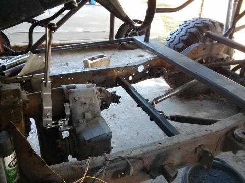 1986 Ford Bronco - The rusty frame of a custom off-road vehicle is shown, with a transfer case and shifter lever prominently visible. A large, knobby tire is…