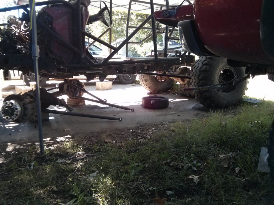 Custom off-road vehicle chassis and drivetrain disassembled, front axle on ground, red truck bed in background.