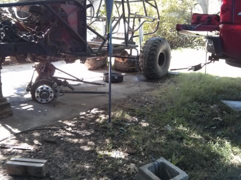 1986 Ford Bronco - A heavily modified off-road vehicle frame is shown with its front wheel assembly detached and resting on the ground. A red pickup truck is…