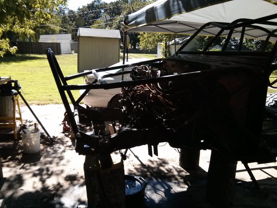 Custom vehicle frame and engine components outdoors under a canopy.