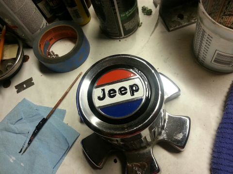 1982 Jeep Wagoneer - A close-up view of a chrome Jeep emblem with red, white, and blue coloring is shown on a workbench. Various automotive repair items like ta…
