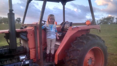 1979 Ford Bronco - A young girl stands on the steps of an orange tractor in a grassy field under a cloudy sky.