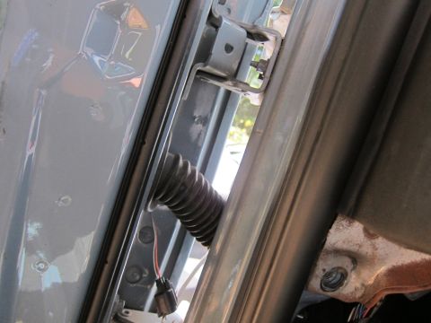 2001 Ford Crown Victoria - A close-up view of the driver's side door jamb area of a light blue vehicle, showing a rubber boot protecting wiring and a door hinge.