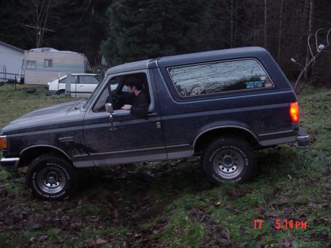 1987 Ford Bronco - A dark blue 1987 Ford Bronco is parked on uneven ground, with a driver visible inside the cabin.