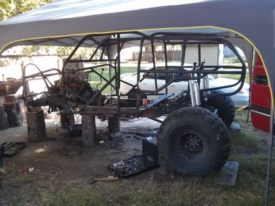 Off-road vehicle chassis with engine and suspension components, supported by logs. Tools are on the ground