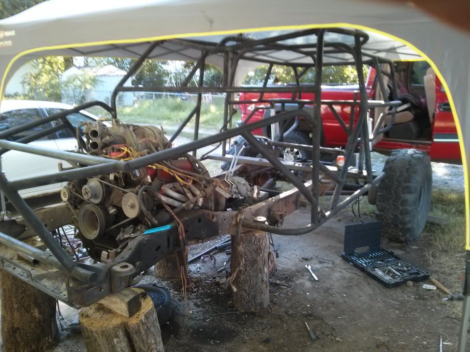 off-road vehicle chassis on blocks, engine exposed, red truck in background