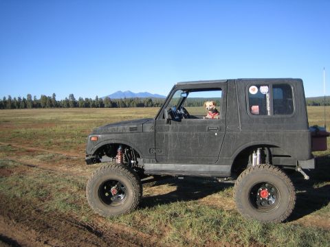 1987 Suzuki Samurai - A dog sits in the driver's seat of a lifted Suzuki Samurai, parked in a field with mountains visible.