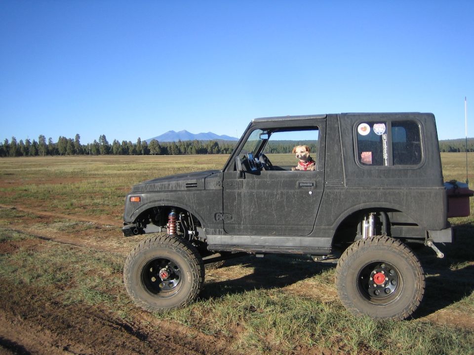 Side view of a lifted 1987 Suzuki Samurai with a dog in the driver's seat, parked in a grassy field with mountains in the background