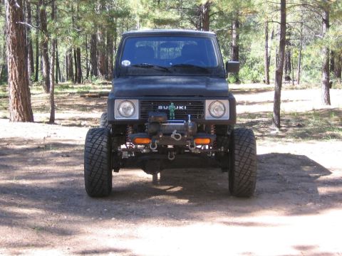 1987 Suzuki Samurai - A black 1987 Suzuki Samurai is parked in a wooded area, showing its front grille and large off-road tires.