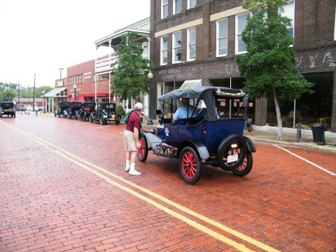 1909 Ford Model T - A line of vintage touring cars, including a 1909 Ford Model T, parked on a historic brick street.