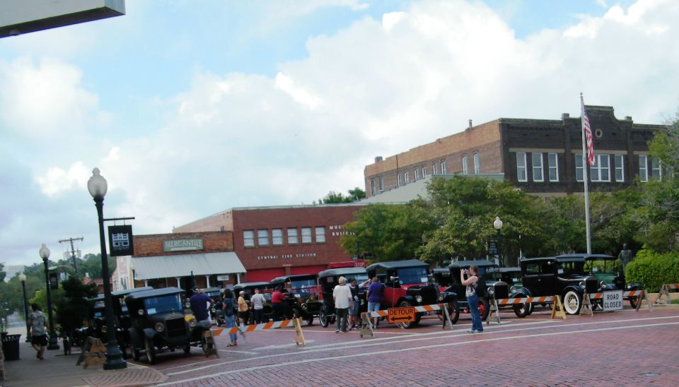 Several old cars are lined up on a brick street in front of buildings. People are standing around them