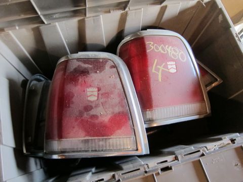 1991 Ford LTD Crown Victoria - Two red taillights with chrome trim are stored in a plastic bin. One has yellow writing and a white emblem.