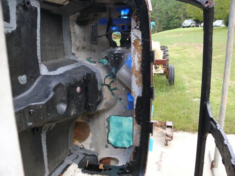 Car interior showing firewall and floor pan during restoration, with a tractor and cars visible in the backgro
