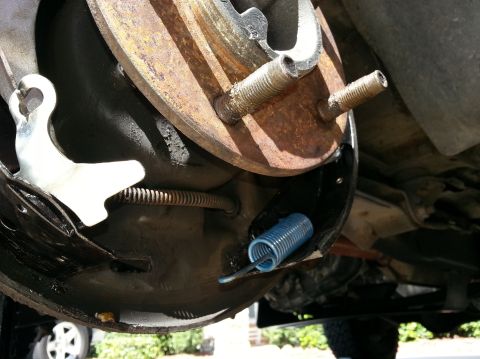 1994 Ford Bronco - Rear drum brake internals viewed from below with blue and bare return springs, rusty wheel studs and brake shoes, vehicle on jack stands.