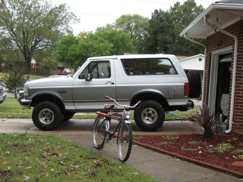 1994 Ford Bronco - A silver 1994 Ford Bronco is parked in a driveway next to a garage, with a vintage bicycle positioned in front of it.