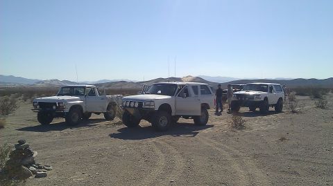 1996 Ford Bronco - A trio of modified off-road vehicles, including a 1996 Ford Bronco, are parked in a desert landscape under a clear blue sky.