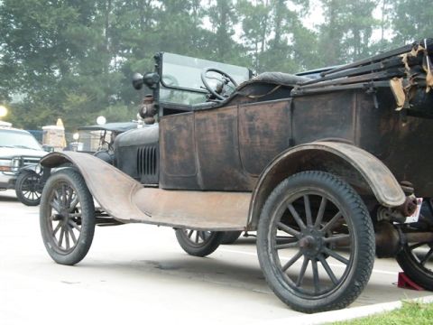 1909 Ford Model T - An old, weathered car sits parked next to another vehicle, with trees in the background.