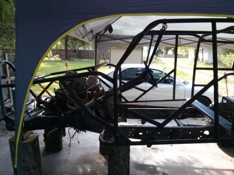 1986 Ford Bronco - A custom-built vehicle chassis with its engine exposed sits under a canopy, partially obscuring a white sedan in-progress car.