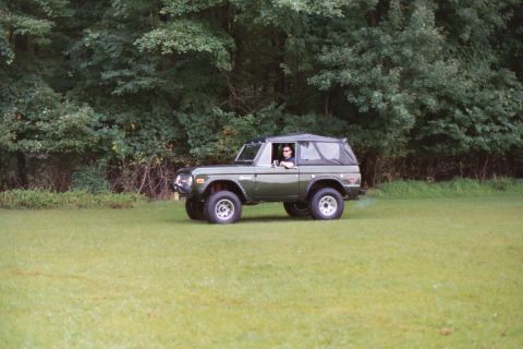 1976 Ford Bronco - A driver is behind the wheel of a green Ford Bronco as it sits on a grassy field with trees behind it.