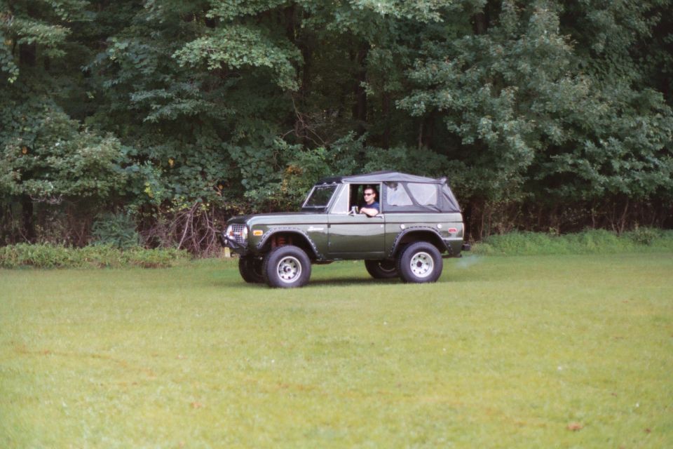 Green 1976 Ford Bronco parked on grass with trees in background. Driver visible