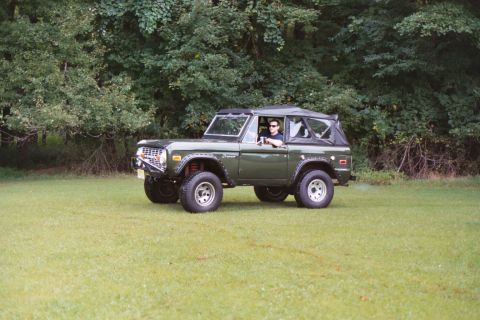 1976 Ford Bronco - A 1976 Ford Bronco is seen from a 3/4 front angle on a grassy field, with a driver behind the wheel.