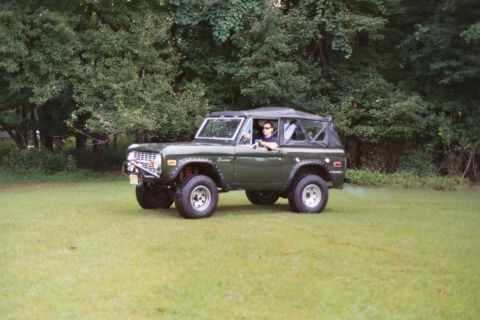 1976 Ford Bronco - A green 1976 Ford Bronco with a soft top is parked on a grassy area with trees in the background.