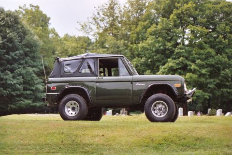 1976 Ford Bronco - A side view of a green 1976 Ford Bronco with a black soft top, positioned on a grassy field.