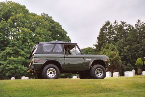 1976 Ford Bronco - A classic green 1976 Ford Bronco is seen from the side, parked in a cemetery with trees and gravestones in the background.