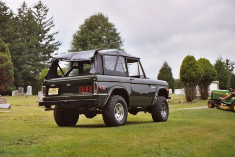 1976 Ford Bronco - A 1976 Ford Bronco is parked on a grassy field near a cemetery, with a riding mower visible to the right.