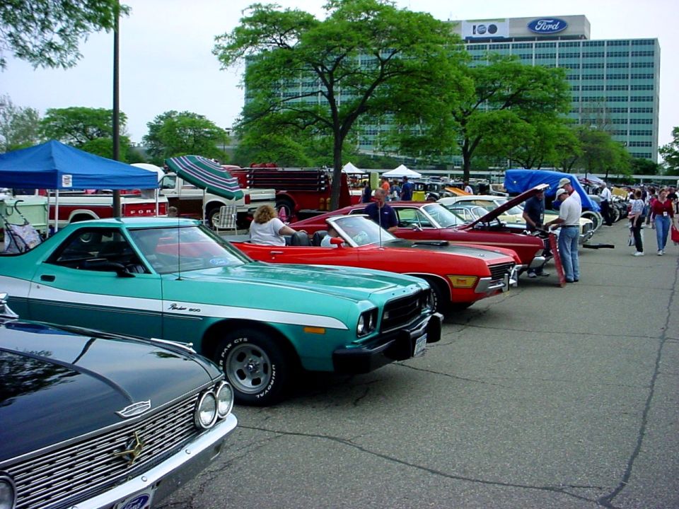 Row of classic cars at a show with Ford headquarters tower behind them