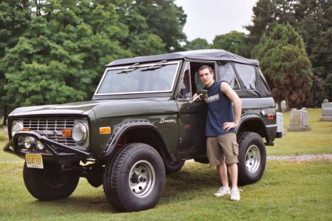1976 Ford Bronco - A man poses with a classic green Ford Bronco in a grassy area with trees and gravestones in the background.