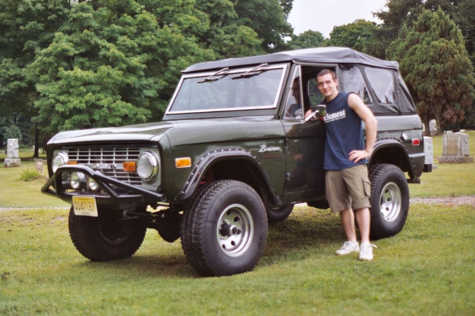 Dark green 1976 Ford Bronco with a soft top and aftermarket bumper, parked on grass with a man standing beside it