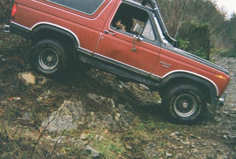 1983 Ford Bronco - Driver-side view of a red 1980s Ford Bronco tilted steeply off-camber on a rocky bank, riding on white-letter General Grabber tires with a…
