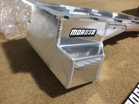 2003 Ford Crown Victoria - Angled view of the deep welded sump end of a fabricated aluminum Moroso oil pan resting on carpet, decal visible at edge.