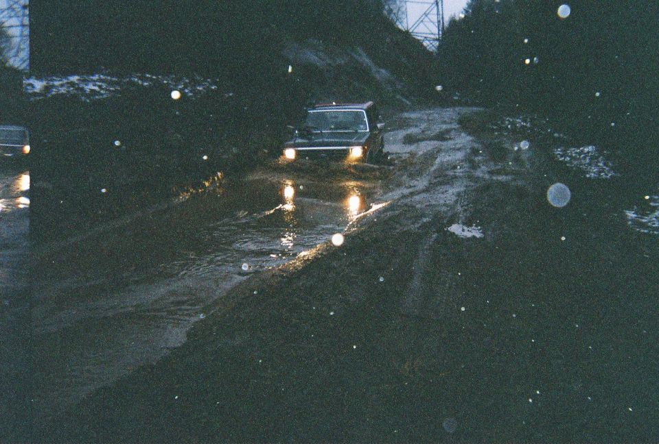 off-road vehicle driving through muddy terrain with headlights on, another vehicle visible to the left