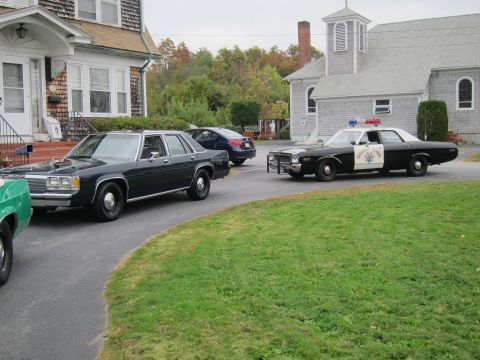 2001 Ford Crown Victoria - A dark sedan and a police car are parked on a driveway in front of a house and a church.