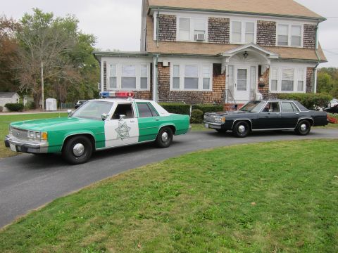 2001 Ford Crown Victoria - Two sedans are parked on a driveway in front of a house. One is a police car.