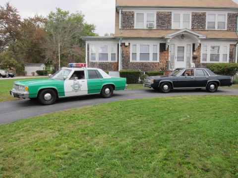 2001 Ford Crown Victoria - Two sedans, one a police car, are parked on a driveway in front of a house.