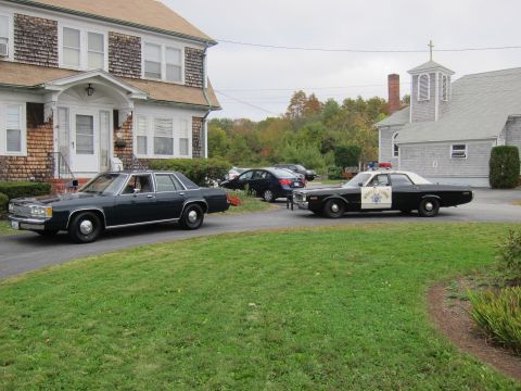2001 Ford Crown Victoria - A 1977 Plymouth Gran Fury police car and a dark sedan are parked on a driveway.